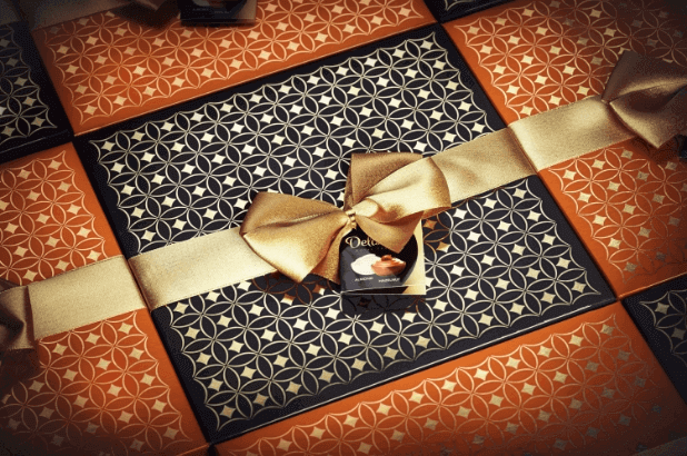 Równo ułożone prezentowe opakowania pralin Delissimo Almond i Hazelnut w eleganckiej, ciemnej i złotej szacie graficznej z dołączoną złotą kokardą